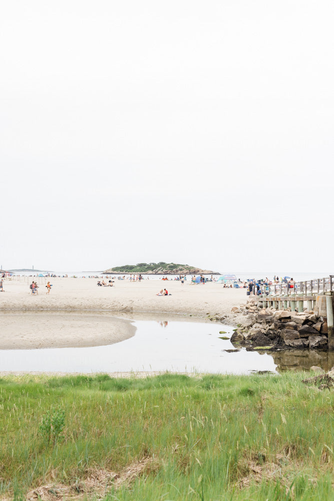 Photograph of a Beach scene at Good Harbor Gloucester with people and a wooden pier, grass in the foreground